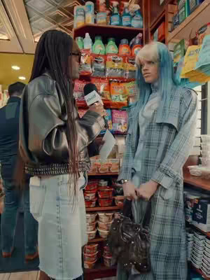 A woman with long braided hair interviews another person who has blue and blonde hair, both are standing in a convenience store setting in front of a packed shelf. The interviewer appears to be in a gray leather jacket, and light-wash denim skirt holding a microphone. The interviewee wears a plaid jacket and holds a designer bag.