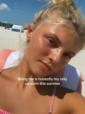 A selfie taken from a slightly high angle shows a young woman at the beach. The woman has blonde hair pulled up in a bun and a light suntan with a bit of shine. She wears a striped bikini top. In the background, a beach setting with the ocean under a blue sky is visible.