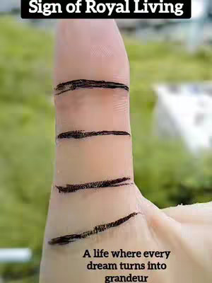 The image shows an up-close view of the side of a hand, with the index finger pointing upwards. Several black lines are drawn horizontally across the fleshy surface of the hand, appearing as markings or symbols. The background is blurred, hinting at a natural outdoor setting, possibly a grassy area.