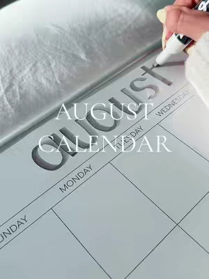 The cover image features a close-up shot of a large paper calendar laying open on a white surface. The word "AUGUST" is prominently displayed in bold, gray, 3D letters. A hand holding a white marker writes on the calendar. The overall composition is clean and minimalist, with a soft, even lighting that adds to the calming aesthetic.