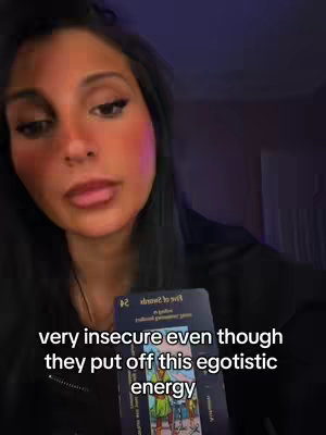 A woman with dark hair is centered in the frame, holding a tarot card. She has a neutral expression and is looking towards the camera. Below her, text overlays read "very insecure even though they put off this egotistic energy". The background is blurred, with purple and pink lighting.