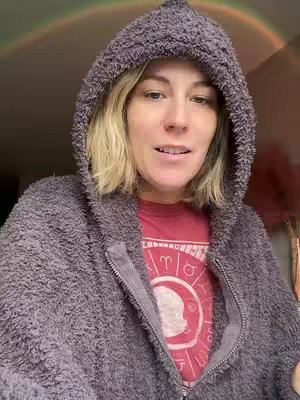 A woman is centered in the frame, wearing a plush gray hoodie, with the hood up. She faces the camera and is wearing a red t-shirt underneath, showing a zodiac design. Natural lighting from behind casts a rainbow in the background. The composition is framed from the mid-chest up, emphasizing the face and upper body.