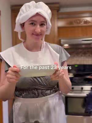 A woman wearing a chef's hat, a white bib, and a grey top holding a white bowl, smiling. The background is a kitchen with wooden cabinets and appliances. Text overlayed on the image.