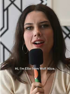 A woman with long dark hair is holding a microphone and speaking. The background is a modern, abstract design in black and white. Text overlay on the image says: Hi, I'm Ellie from Wolf Alice,