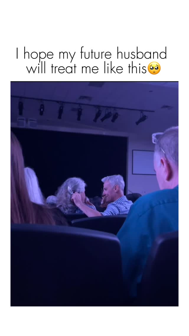 The cover image shows a scene from a dimly lit auditorium, with mostly blue lighting. In the foreground, partial figures of individuals are seen seated. The central focus is a mature couple, with the man in a checkered shirt and the woman with curly gray hair, sharing a tender moment. Above them, text overlays the scene.