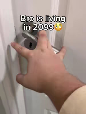 A person's hand reaching to open a door. The hand is positioned near the door handle, which is silver-colored and has a keyhole. The wall is white.