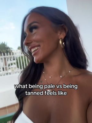 A young woman with long, dark hair and gold hoop earrings smiles broadly in an outdoor setting. Text overlayed says "what being pale vs being tanned feels like". She wears a white top showing a bit of cleavage. The background is blurred, with hints of green trees and blue sky.