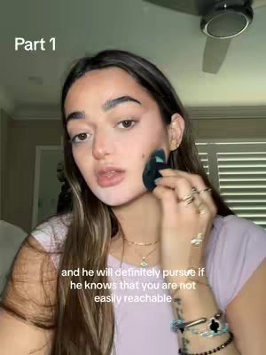 A woman applies makeup with a small sponge, looking directly at the camera. The background is blurred but appears to be a room with natural light from a window and a ceiling fan. Text overlay includes 'Part 1'.