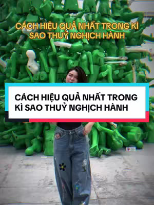 A woman stands in a large room, partially obscured by a sculpture made of green, mannequin-like figures. She is wearing a black top and blue jeans with floral patches. Overlaid text reads "CÁCH HIỆU QUẢ NHẤT TRONG KÌ SAO THỦY NGHỊCH HÀNH" (Most Effective Way in Mercury Retrograde). The text is also repeated in a colored bar across the image's background.