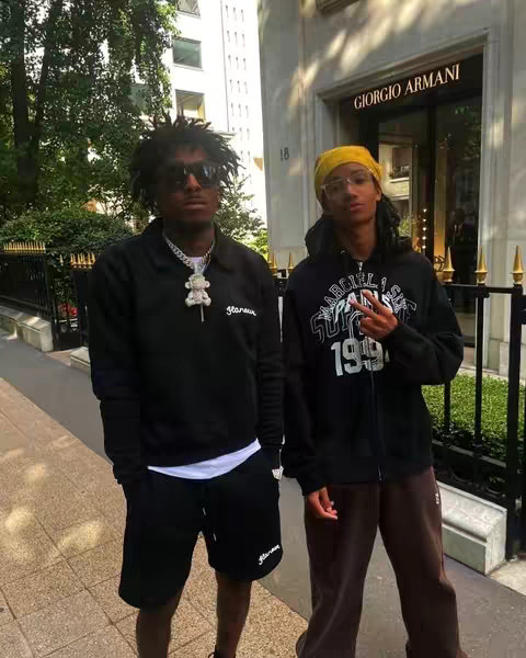 Two young men are standing in front of a store in Paris. One is wearing black and white attire with sunglasses, the other is wearing a hoodie and glasses. The background features a building with the "Giorgio Armani" store sign.