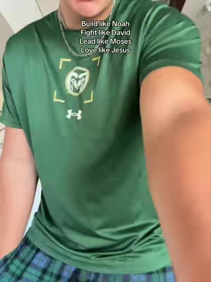 A person is filming with a phone. The person wears a green shirt and has text overlay on top of the image. The background is plain.