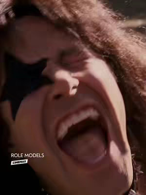 A close-up of a man with wild, brown hair and a black star-shaped eye covering screaming with their mouth wide open. The man appears to be making a comedic face. In the bottom left corner, text reads "ROLE MODELS" and "CINEMAX".