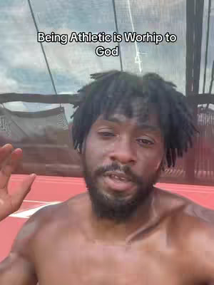 A man with dark skin and short dreadlocks is centered in the frame, shirtless and looking directly at the camera. He is standing in what appears to be an athletic field or track setting, with red track lines visible in the background and a partial view of a blue sky and netting overhead. The man has a beard and appears to be sweating. Text overlayed at the top reads 'Being Athletic is Worship to God.'