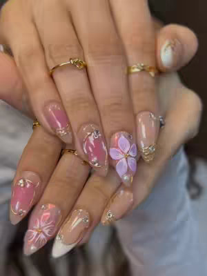 A close-up view of a hand holding a well-manicured hand with long, pointed nails. The nails are adorned with a mix of pink, nude, white, and floral designs, set against a soft, blurred background. Gold rings decorate various fingers, enhancing the overall elegant aesthetic.