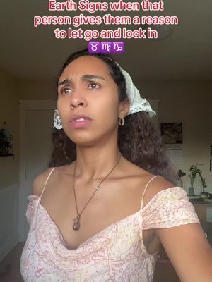 A young woman looks up with a thoughtful expression, her face framed by dark curly hair and a white lace headscarf. She wears a light, patterned dress and a pendant necklace. Text overlays at the top of the image read 'Earth Signs when that person gives them a reason to let go and lock in.' There is a light-colored wall behind her.