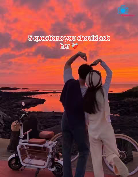 A couple stands embracing with arms raised to form a heart shape against a vivid sunset. A scooter is parked in the foreground, and the sea is visible in the background, adding to the romantic ambiance.