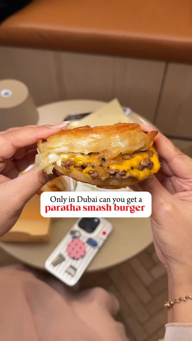 A person's hands are holding a half-eaten smash burger in a paratha bun, with a background that is partially visible showcasing of a wooden table and other food items.