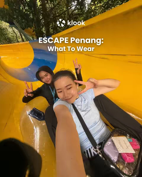 Two women smiling and giving peace signs while sitting on a yellow water slide. The slide is in the background, and they appear to be at a water park.