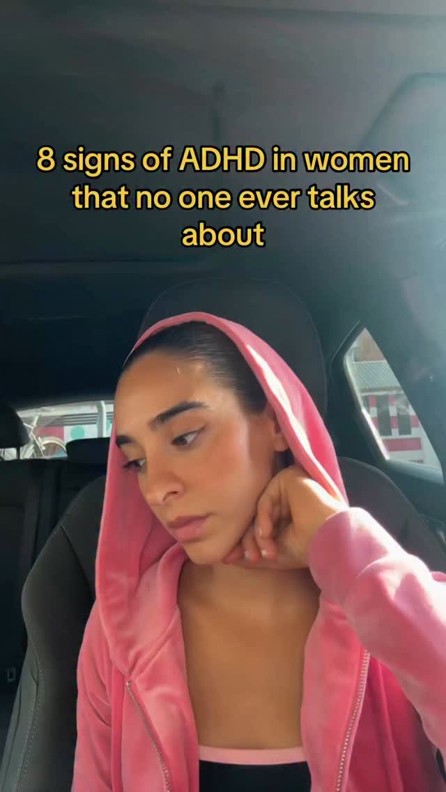 A woman with dark hair is sitting in a car, looking down with a contemplative expression. She is wearing a pink hoodie, and the interior of the car is visible in the background. Text overlay reads: '8 signs of ADHD in women that no one ever talks about.'