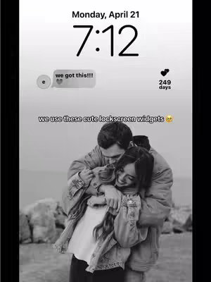 A black and white photo of a couple hugging in front of a blurred outdoor background. The focus is on the couple, with the man embracing the woman from behind. The image has a romantic, intimate feel.
