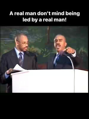 The cover image features two men in suits. One, on the left, is wearing glasses and looking towards the other man, who has his right arm raised. The second man is pointing his right index finger toward the viewer and holding a microphone. Text in white letters is at the top. The background is blurred, with muted colors.
