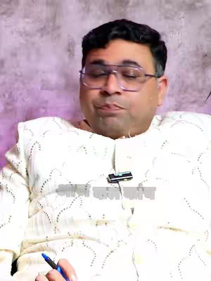 A man with closed eyes and glasses sits in front of a purple-toned background. He wears a white, intricately patterned shirt. A microphone is clipped to his shirt, just below the collar. Below the man and his shirt is a logo in Hindi. The man holds a blue pen.