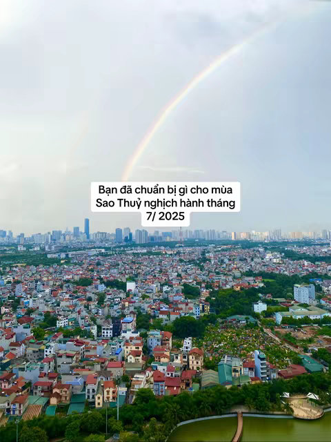 An aerial view captures a cityscape under a sky filled with clouds and a vibrant rainbow arcing across the frame. The city features a dense collection of buildings with diverse roof colors, suggesting residential areas. The layout gives a sense of urban sprawl, fading into the distance. In between the buildings and the city there is a green space with water, the scene is framed by a cloudy sky and a full rainbow centered above the city.