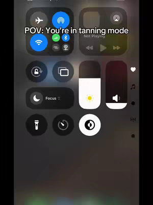 The image displays a phone's control center interface, featuring various icons and controls against a blurred, dark background. At the top, the text "POV: You're in tanning mode" overlays the screen. The control center includes icons for Wi-Fi, Bluetooth, music controls, and a slider for screen brightness set to display the sun.