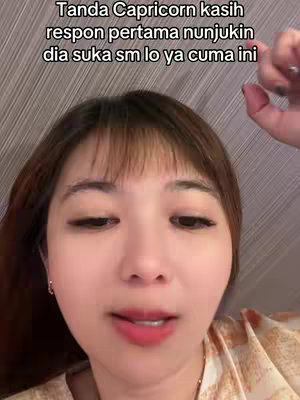 A close-up shot features a woman with fair skin and dark brown hair with bangs, looking slightly towards the left. The background is a neutral beige color, and she is wearing a patterned shirt. Above her head is text, and she’s holding her right hand up near her head.