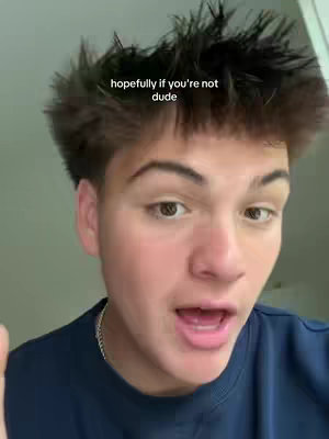 A medium shot of a young man's face, with the upper half of his head and shoulders visible. He has short, dark spiky hair with a skin fade. The man is looking directly at the camera with his mouth open. Overlaid text reads: 'hopefully if you're not dude'. He is wearing a dark blue shirt. The lighting is somewhat soft and diffuse.