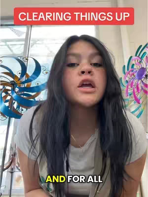 A young woman with long dark hair is centered in the frame, looking directly at the viewer and speaking. The background includes a window and colorful decorative pinwheels. Text overlays at the top and bottom of the image read, "CLEARING THINGS UP" and "AND FOR ALL". The overall composition is a selfie-style shot with eye-level framing.