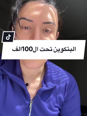 A woman with dark hair and eyes is centered in the frame, with the upper half of her body visible. She's wearing a blue athletic zip-up jacket and a necklace. A white rectangular overlay with the text "البِتكوين تحت ال 100 ألف" (Bitcoin below 100K) is placed at her chest. The background is slightly out of focus, and the lighting suggests it's indoors.