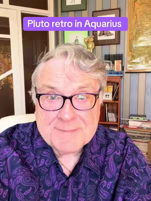 A man with gray hair and glasses smiles at the camera. He is wearing a patterned purple shirt. Above him is a rectangular text overlay that reads "Pluto retro in Aquarius". The background includes a wall with paintings, a wooden cabinet, and a window with curtains.