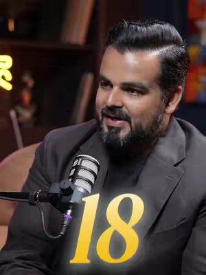 A man with a beard and dark hair, seated and speaking. He wears a dark blazer over a dark t-shirt. Behind him, out-of-focus are bookshelves. Superimposed on his chest is a yellow number '18' with a glowing effect.