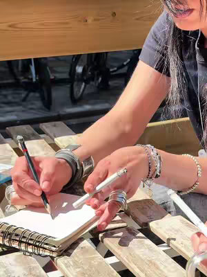 Close-up shot of a person sketching in a notebook while seated at a wooden table outdoors. The hands, adorned with multiple rings and bracelets, hold a pencil and a pen. The background blurred, showing bicycles under a wooden structure, and bright sunlight.