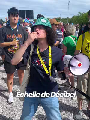 A crowd of people gathered outdoors with a man holding a megaphone in the foreground. The man, wearing a patterned hat and a lanyard, is mid-action with the megaphone near his mouth. Text overlayed at the bottom reads 'Échelle de Décibel'. People are blurred in the background.