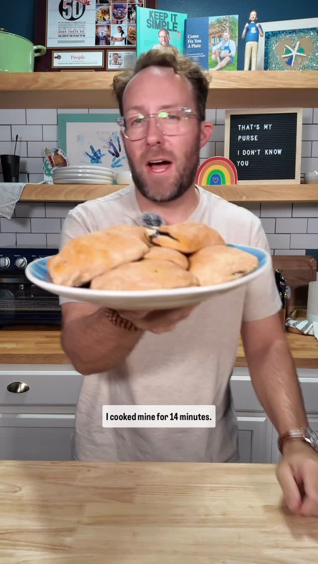 A man stands in a kitchen, holding a plate of blueberry pies. Behind him are shelves with a collection of items, including books and a sign. The man is looking at the camera and talking.