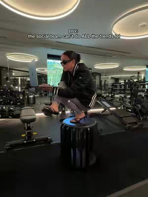 A woman wearing sunglasses and heels is sitting on a cylindrical platform with a laptop in her lap. She is in a gym, and there is equipment in the background.