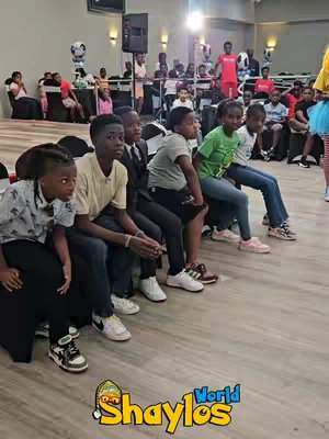 A group of children and young adults are seated, some in chairs, others on a bench. The setup suggests an event or party. In the foreground, several children are seated on the bench, appearing to be listening to the event happening, the background consists of a venue decorated with what appears to be balloons.