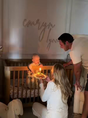 A cozy indoor scene, featuring a baby in a wooden crib. The baby is focused on a birthday cake with a lit candle. The mother is holding the plate and is looking at the baby, while the father watches from outside.