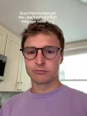A man with brown hair and glasses looks at the camera with a slightly inquisitive and serious expression. The background is of a kitchen setting with white cabinets.