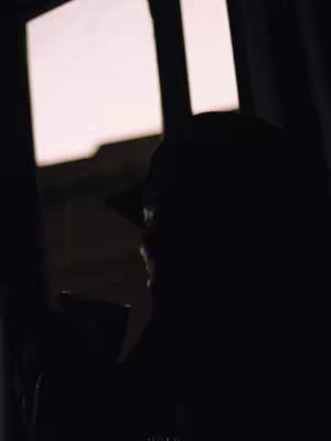 A silhouette of a person wearing a cap, viewed from the side, is positioned in front of a window with bright light flooding in. Dark interior details include a table or ledge.