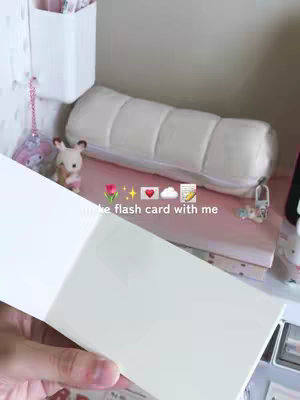 A hand holding open a notebook or note cards with a desk and various storage and decor items in the background.