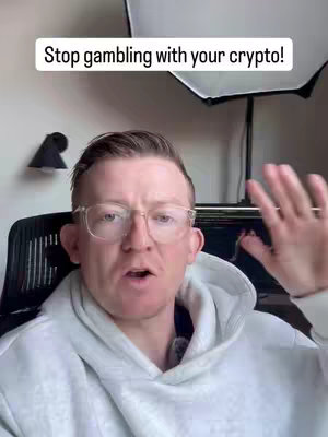 A man with glasses and a white hoodie is gesturing with his right hand while looking directly at the camera. Text at the top reads, 'Stop gambling with your crypto!' The background appears to be a room with a lamp and a light panel.