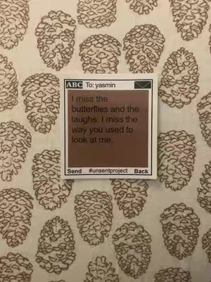 The image captures a close-up of a piece of paper with a handwritten message against a backdrop of fabric patterned with brown, stylized pinecone shapes. The paper is a rectangular sticky note-like frame, its central area colored a solid brown, enclosed by a white border. Within the brown area, the message is written in black text. At the top left of the note, there is a grey rectangle, and the words 'To: yasmin' are visible beneath the box. The message on the note contains heartfelt words: 'I miss the butterflies and the laughs. I miss the way you used to look at me.' It appears the message is formatted like a text with the send, unsentproject and back as buttons at the bottom, the background of the image is a fabric with a pattern of beige pinecones scattered across its face.