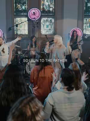 The cover image shows a live performance scene in a church setting. Performers, including a lead singer with blonde hair, are on a stage in front of a drum kit and other musical instruments. The audience is partially visible, and the background features stained-glass windows and spotlights in the periphery, with stage lighting that enhances the scene and highlights the performers. Text 'SO I RUN TO THE FATHER' is added to the image.