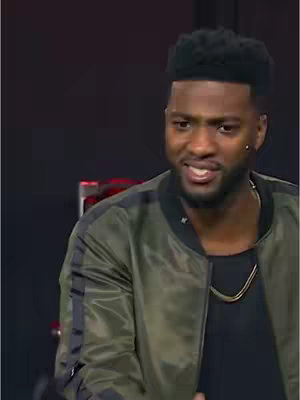 A man with a beard and short, dark, curly hair is seen facing the camera in a medium shot. He is wearing a green and black bomber jacket over a black shirt and a gold chain. The background appears to be a dark setting with some indistinct shapes and shadows.