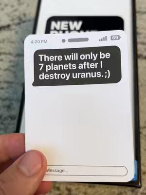 A hand holds a card-shaped object resembling a phone displaying a message. The background is dark, while the object is predominantly white. The text on the screen reads, 'There will only be 7 planets after I destroy uranus. ;)' It also features the time on the top-left, the battery level, and service bars on the top-right and a message area at the bottom.