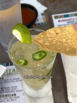 A close-up shot showing a cocktail in a clear glass. The drink is a light, yellowish color, and it contains pieces of green jalapeño and a lime wedge. A tortilla chip is held in the frame, about to dip into the drink. The background is blurred, suggesting the photo was taken at a restaurant or bar, with text on a folded paper.
