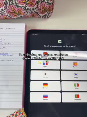 The image shows an iPad displaying a language learning app's interface and handwritten notes to the left. Several language flags are visible on the app's screen. On the top, a printed fabric lies, set on a white surface.